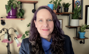 A blue-eyed, white person in the foreground with long, curly hair and smile on her face. In the background, plants are on wall shelves.