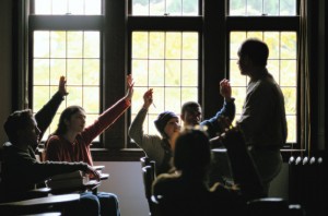 Several college students in a classroom with a professor. All of the students have their hands raised.