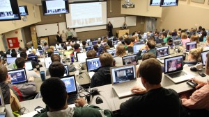 This is a picture of dozens of students in a large lecture hall. All of the students have computers out in front of them and there are large screens around the whole room.